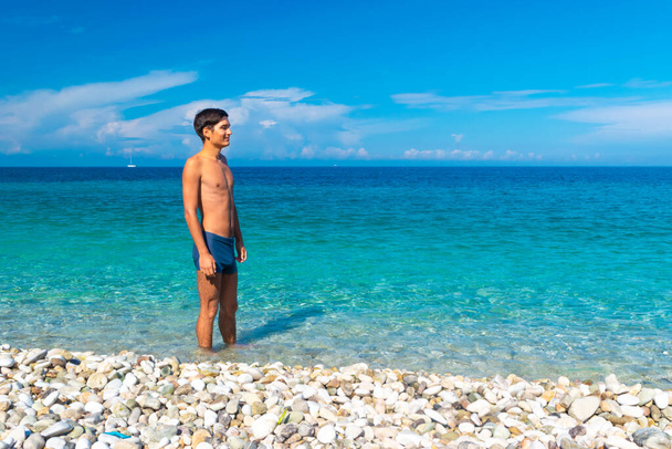 Die Figur eines jungen, braun gebrannten Mannes am Strand vor dem Hintergrund des blauen Meeres und Himmels. Das Konzept eines Sommerurlaubs am Meer, Vitamin D beim morgendlichen Sonnenbad zu bekommen. - Foto, Bild