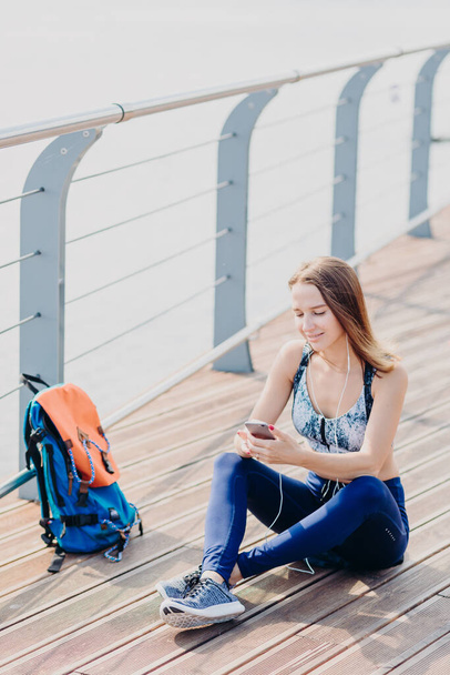 Outdoor shot of pleasant looking relaxed woman sits crossed legs, looks positively at smart phone, listens music with earphones enjoys high speed internet. Female traveller takes break after long walk - Photo, image