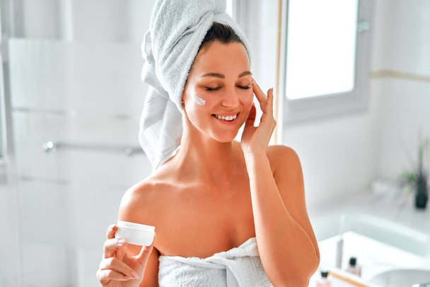 Bella donna in un asciugamano bianco in piedi al lavabo e specchio con una crema idratante per il viso. routine di cura della pelle mattina. Cosmetologia e procedure termali. - Foto, immagini