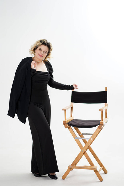 Una mujer sonriente con un traje negro se apoya sobre una silla sobre un fondo blanco. - Foto, imagen