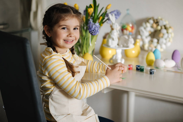 Söpö neljän vuoden tyttö beige esiliina istuu keittiön pöydällä ja maalaus pääsiäismunia. Laadukas kuva - Valokuva, kuva