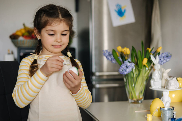 Feliz niña pintando huevos de Pascua en la cocina. Linda chica sonriente divertirse en vacaciones de primavera. Foto de alta calidad - Foto, imagen