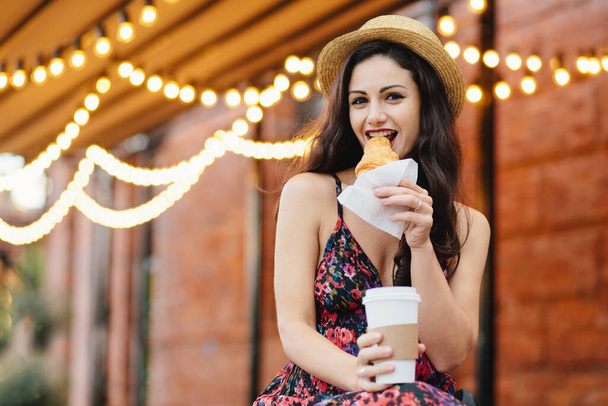 Pretty woman with dark hair, shining eyes eatting with pleasure tasty croissant and drinking takeaway coffee while sitting at cozy balcony. Young woman in summer hat and dress relaxing at cafe - Foto, Imagem
