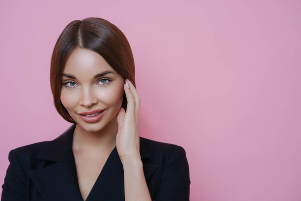 Photo of lovely European female entrepreneur with healthy skin, touches face gently, returns from spa salon, wears formal black outfit, looks confidently at camera, isolated on pink background - Foto, imagen