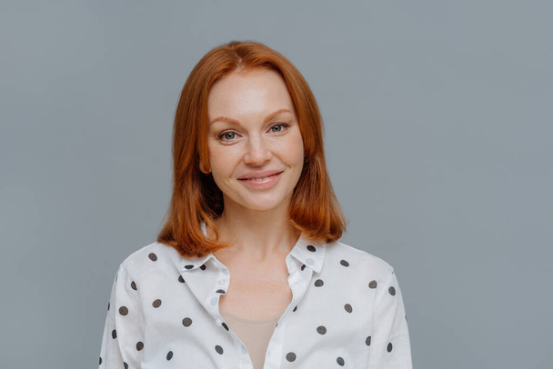 Portrait of attractive ginger European woman smiles gently at camera, wears makeup, dressed in polka dot blouse, looks directly at camera, models against grey background, thinks about changing job - Photo, Image