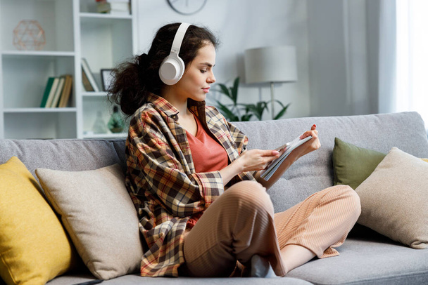 Curly-haired gir studentl sits on the sofa, wears headphones, looks in notebook and checks notifications on the phone. A beautiful woman studies and switches the music that will play. - Foto, immagini