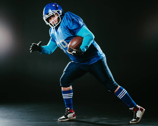 Retrato de un hombre con un uniforme azul para carreras de fútbol americano con una pelota sobre un fondo negro - Foto, imagen