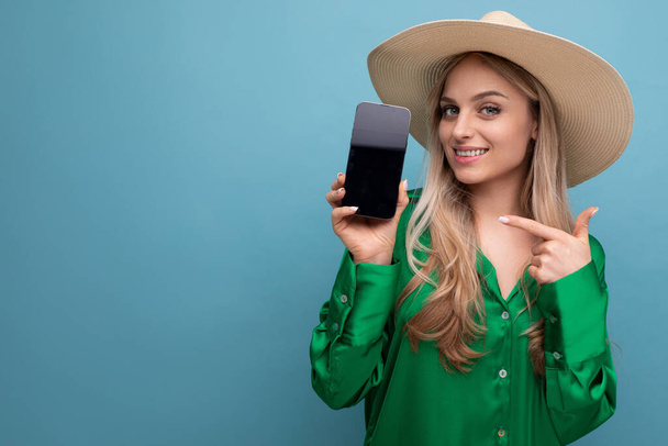 słodkie młoda kobieta na wakacjach mając na sobie letni słomkowy kapelusz pokazując ekran telefonu z mocup na niebieskim tle studio. - Zdjęcie, obraz