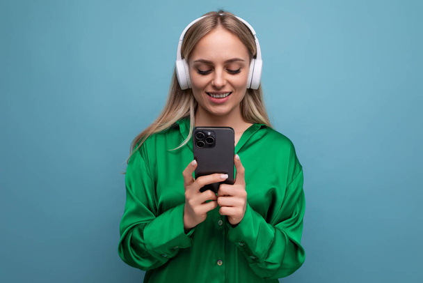 foto de una linda joven rubia atractiva en auriculares con un teléfono inteligente en sus manos sobre un fondo azul. - Foto, Imagen