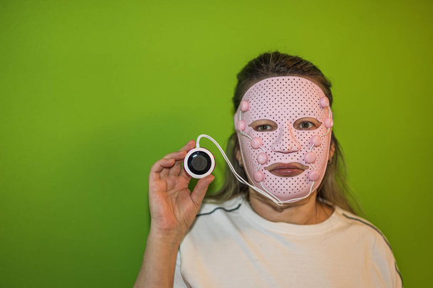 Közelkép egy rózsaszín arcmasszírozó maszkos nőről, aki távirányítót tart kijelzővel. Svédország.  - Fotó, kép
