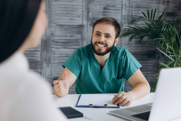 Χαμογελώντας νεαρός καυκάσιος άνδρας γιατρός με ιατρική στολή συμβουλεύεται γυναίκα ασθενή. - Φωτογραφία, εικόνα