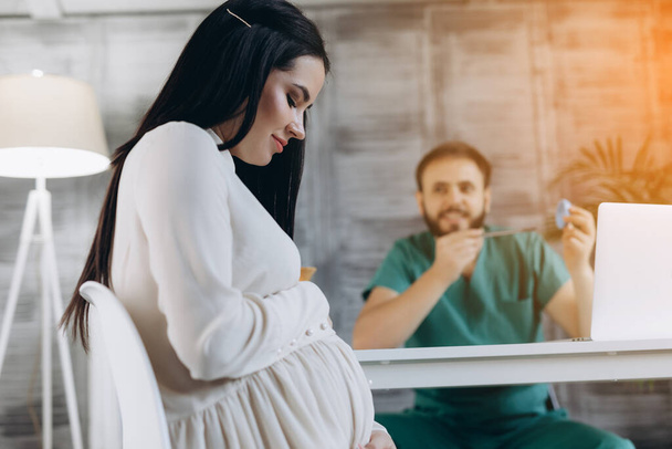 Zwangere vrouw op bezoek bij een arts in de kliniek. Aantrekkelijke vrouw met gynaecoloog op achtergrond. - Foto, afbeelding