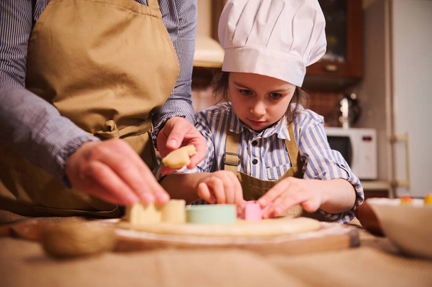 Селективный акцент на вдохновленную маленькую девочку в шляпе шеф-повара готовит пряничное печенье, режет тесто с помощью кусачки печенья, под руководством ее мамы на домашней кухне. Пасхальная концепция - Фото, изображение