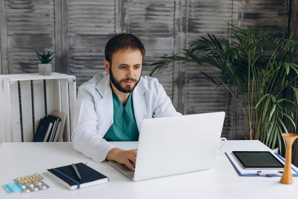 Attraente giovane medico maschio sta lavorando con il computer portatile in armadietto leggero. Sorridendo e guardando la macchina fotografica. - Foto, immagini
