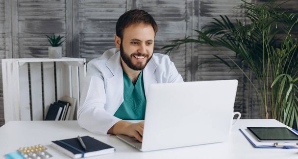 Atrakcyjny młody lekarz pracuje z laptopem w jasnej szafce. Uśmiechając się i patrząc w kamerę. - Zdjęcie, obraz