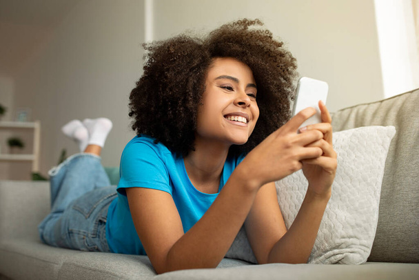Wesoła młoda, kręcona, afrykańska kobieta pisząca na smartfonie, ciesząca się sieciami społecznościowymi, rozmawiająca, leży na kanapie we wnętrzu salonu. Aplikacja i gadżet do surfowania po Internecie, grać w grę w domu - Zdjęcie, obraz