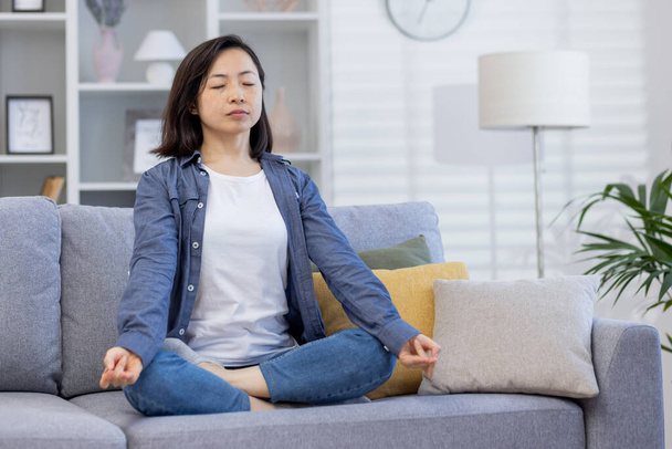 Young beautiful Asian woman alone at home, meditating sitting on sofa in living room in lotus position, sitting with eyes closed at home. - Foto, Bild