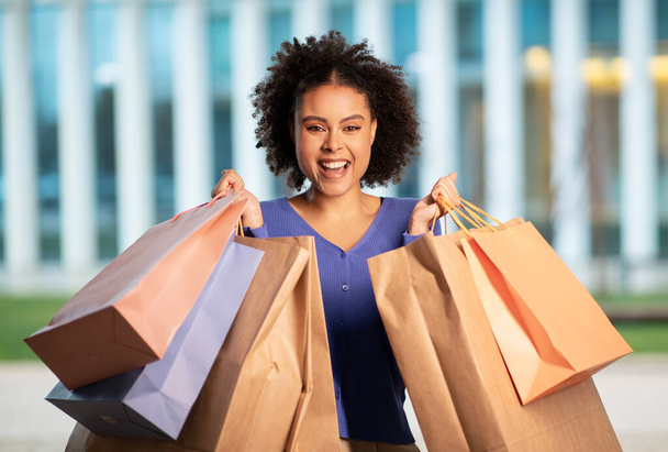 Radosna czarna kobieta trzymająca papierowe torby na zakupy uśmiechnięta do kamery stojącej na zewnątrz w obszarze miejskim. Kobieta Kupujący Zakupy Cieszący Sezon Sprzedaży W Weekend. Koncepcja konsumencka - Zdjęcie, obraz