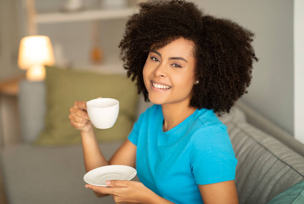 Cheerful pretty young curly african american lady with cup of hot drink, sits on sofa, enjoys relaxing in living room interior. Rest with tea, coffee break at home in free time - Фото, зображення
