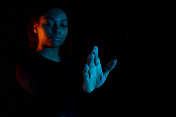 Sanal gerçeklik kavramı. Genç Afrikalı Amerikalı kadın elini uzatıyor ve görünmez dokunmatik ekrana dokunuyor, çok renkli neon ışığı olan koyu bir arka plan, serbest alan. - Fotoğraf, Görsel
