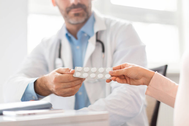 Middle aged male doctor giving pregnant lady prescribed pills blister pack, sitting at table in clinic office, selective focus on hands holding tablets - Photo, image