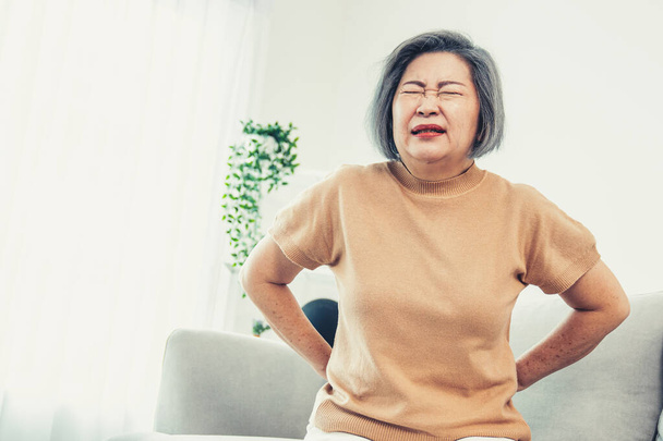 An agonizing senior woman experiencing back pain while sitting on her sofa at home and holding her back. - Foto, Imagem