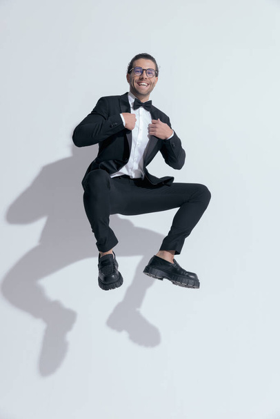 sexy businessman jumping and squatting while fixing his tux, wearing glasses against white studio background - Photo, Image