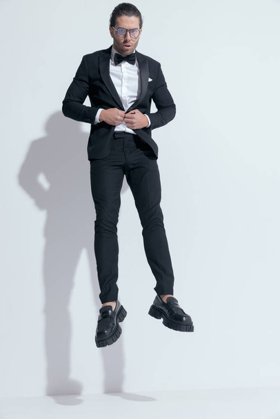 attractive businessman with tough vibe jumping and closing his tux, wearing glasses against white studio background - Foto, immagini