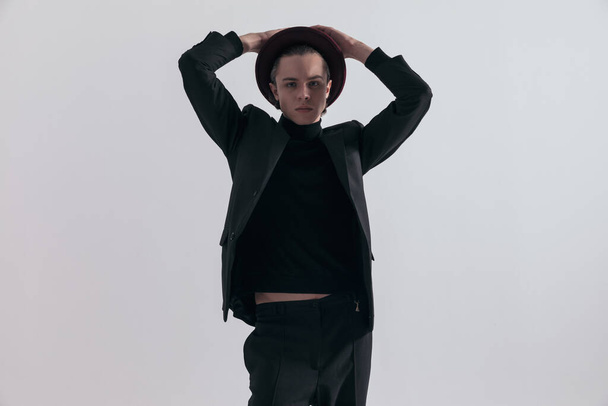 Fashion picture of young businessman arranging his hat and wearing a stylish outfit against gray studio background - 写真・画像