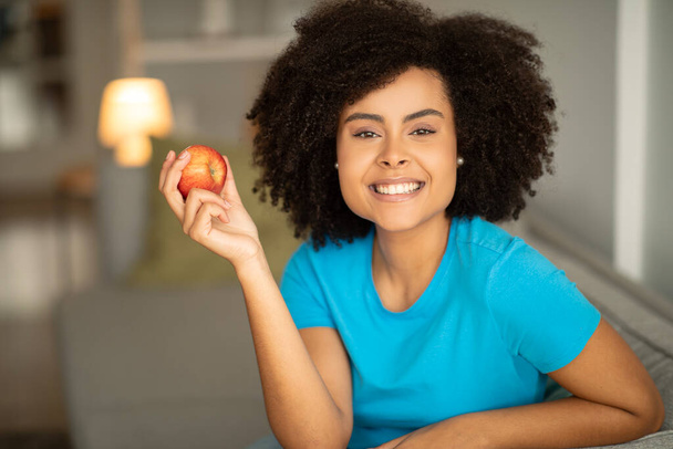 Smiling young curly african american woman with apple sits on sofa, enjoys relaxing in living room interior. Health and teeth care, useful food, snack and relax at home in free time - Foto, Imagem