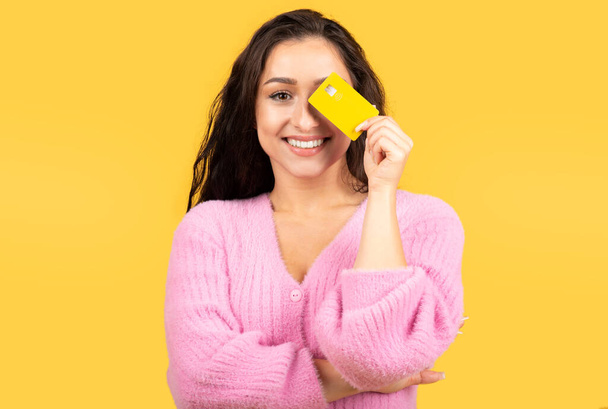 Cheerful young caucasian brunette lady put credit card to eye, has fun, isolated on yellow studio background. Huge sale, discount, banking, finance saving, money and online shopping - Foto, Bild