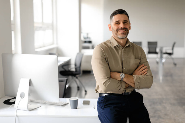Successful middle aged male entrepreneur sitting with arms crossed at desk in office, looking and smiling at camera, free space. Confident male graphic designer working at company - Photo, Image