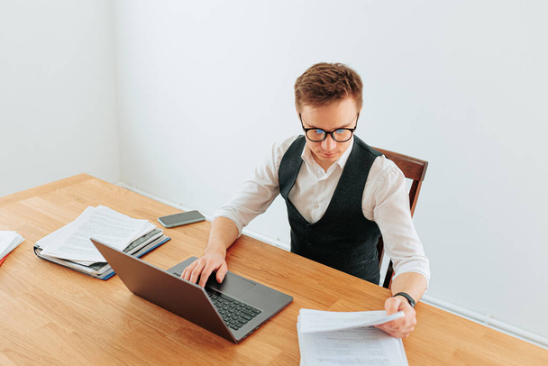 A young professional in his home office, dressed in a white-collar shirt and glasses, types on his laptop, surrounded by books and papers, with a city view in the background. - Foto, afbeelding