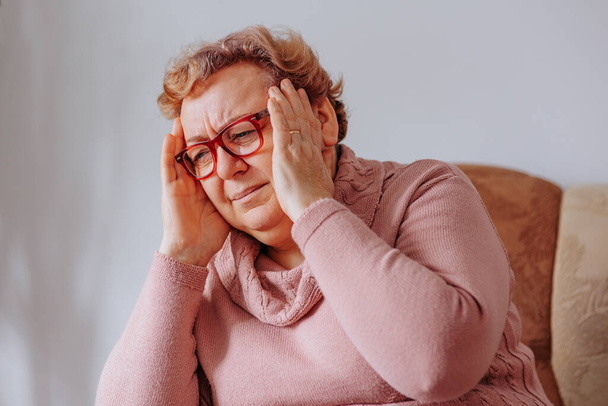Senior woman grimacing in pain, with her hands on her head, experiencing a throbbing headache at home, Coping with Headaches Senior Woman Finds Support at Home - Fotoğraf, Görsel