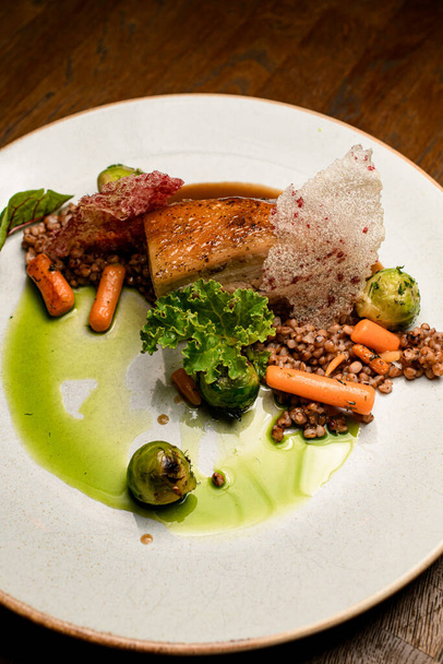 view on plate with dish of piece of baked pork meat with layers of bacon and skin with buckwheat and vegetables and sauce - Фото, зображення