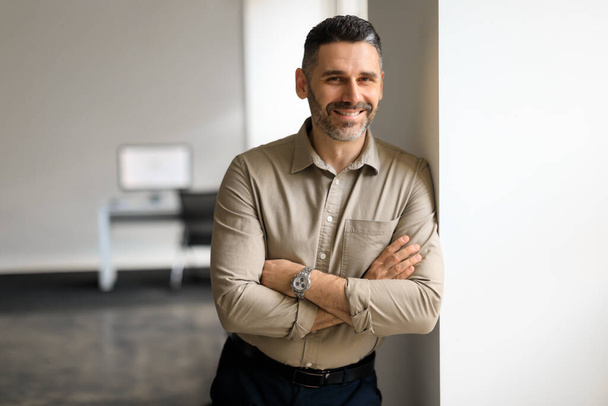 Portrait of confident middle aged businessman in shirt standing in office hall, posing with crossed hands and leaning on wall, looking and smiling at camera, copy space - Фото, зображення