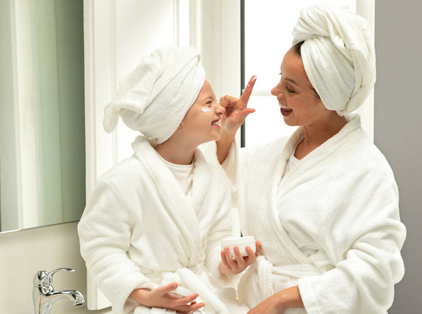 バスローブとタオルで幸せな白人のティーンと若いお母さんは、楽しみを持って、クリームを適用し、バスルームのインテリアで美容ケアをお楽しみください。毎日の手順,自宅での関係,皮膚の水分補給 - 写真・画像