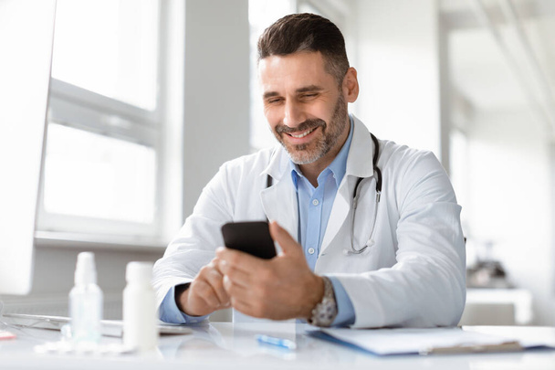 Hymyilevä mies lääkäri valkoisessa lääkärin univormussa istuu työpöydän ääressä klinikalla, katselee kännykän näyttöä, konsultoi asiakasta verkossa. Happy Man GP tai lääkäri älypuhelimella - Valokuva, kuva