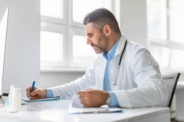 Middle aged male doctor sitting at table in office hospital, therapist man practitioner writing and working in clinic, checking patients results and writing prescriptions - Photo, Image