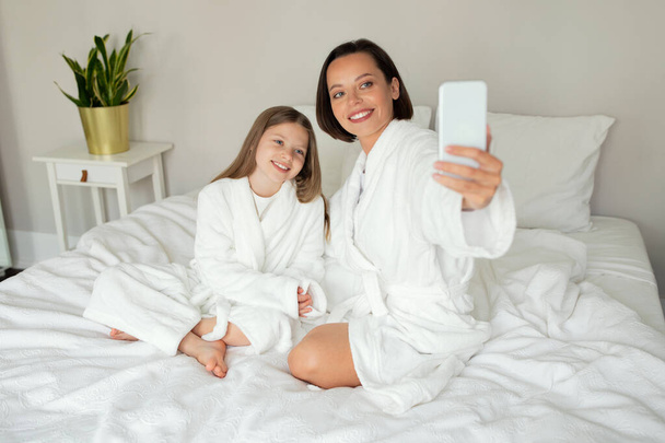 Glad caucasian young lady, teen girl in bathrobe take selfie on phone, shoot video for blog, have video call, enjoy relax on white bed in bedroom interior. Mother daughter relationship at home - Foto, Bild