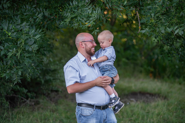 Bald man in glasses holds child in arms in the park. Father in jeans hugs son in nature outside the city. Little boy laughs, having fun with dad on a journey in the forest. Children Protection Day - Valokuva, kuva