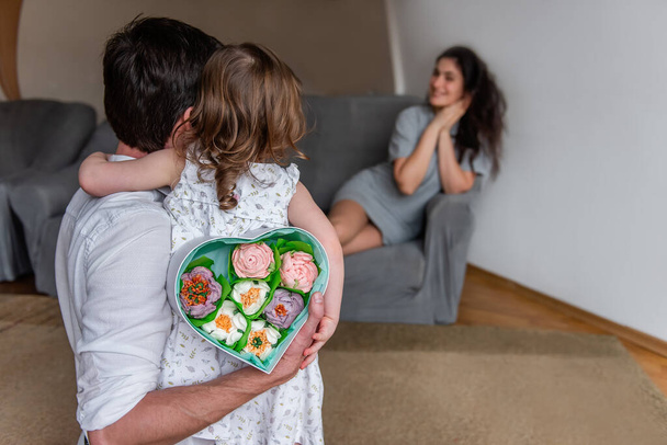Little daughter and father hold gift for mother behind backs, heart shaped box with homemade marshmallow flowers. Surprise for woman on mothers day from child and husband. Girl is emotionally happy - Valokuva, kuva