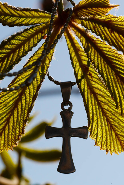 Wisiorek z krzyżem na młodym zielonym liściu kasztanowca wiosną. Symbol religijny. - Zdjęcie, obraz
