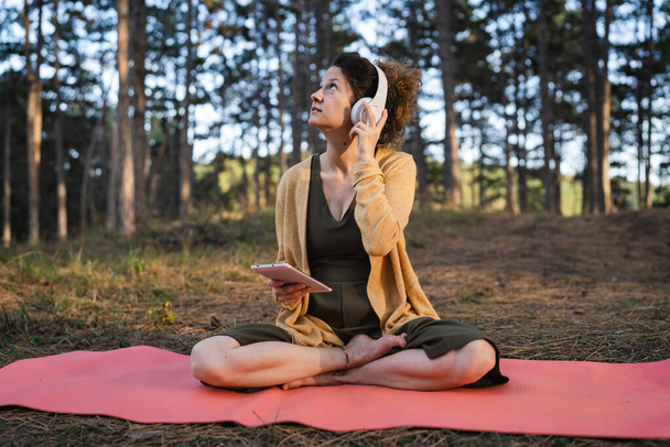 Een vrouw jong volwassen blanke vrouw zit op de yoga mat alleen in het park of bos in de natuur met digitale tablet en het aanpassen van koptelefoon voor te bereiden op online begeleide meditatie zelfzorg - Foto, afbeelding