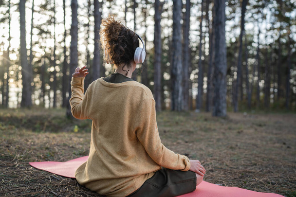 Een vrouw jong volwassen blanke vrouw zit alleen in het park of bos in de natuur met koptelefoon voor online geleide meditatie zelfzorg mentaal evenwicht en manifestatie praktijken concept - Foto, afbeelding