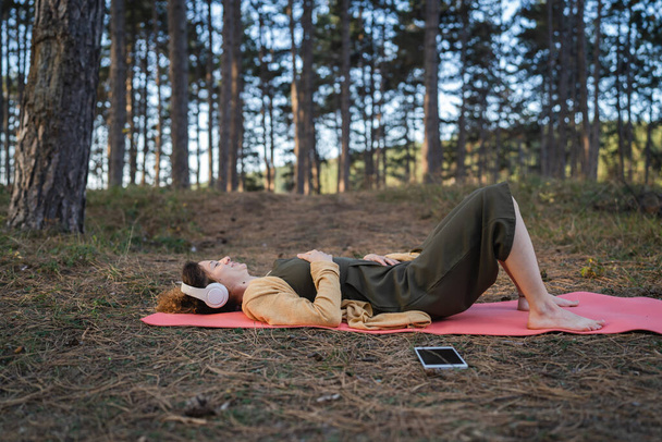 公園や森の中に一人で横たわっている一人の女性の若い大人の白人女性は、オンラインガイド付き瞑想セルフケア精神的なバランスと症状の実践の概念のためのヘッドフォンで自然 - 写真・画像