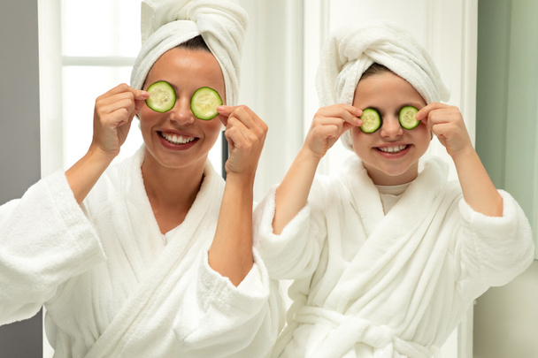 Smiling caucasian little girl and young woman in bathrobe and towel have fun, put cucumber to eyes, enjoy spare time and beauty care in bathroom. Relationship mom and daughter at home - Photo, image