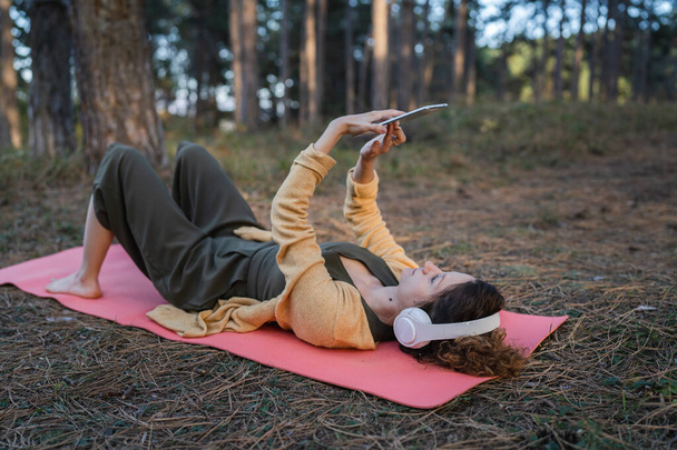 Een vrouw jong volwassen blanke vrouw ligt alleen in het park of bos in de natuur met koptelefoon en digitale tablet voor online begeleide meditatie zelfzorg mentaal evenwicht manifestatie praktijken concept - Foto, afbeelding