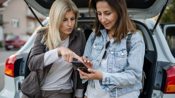 Two women mature female caucasian friends checking mobile phone vacation concept online map planning travel route at car automobile in day real people copy space smart-phone app and GPS - Valokuva, kuva
