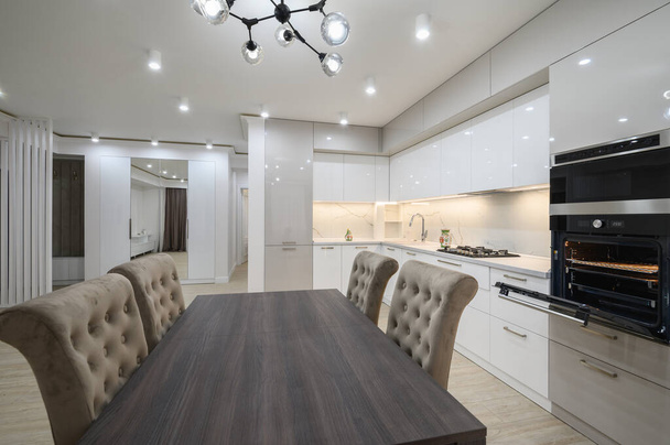 Dining room table and chairs, white spacious kitchen interior with television on the wall in the background and an oven with door open - Photo, Image